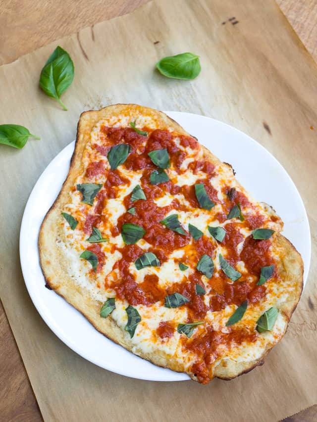 A homemade pizza Margherita on a white plate next to basil leaves