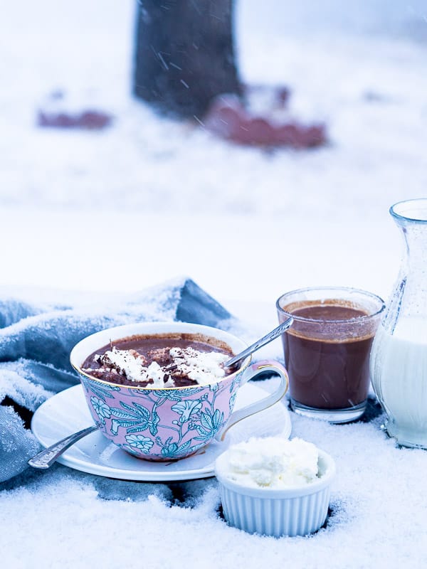2 cups of hot chocolate next to containers of whipped cream and milk in front of falling snow 