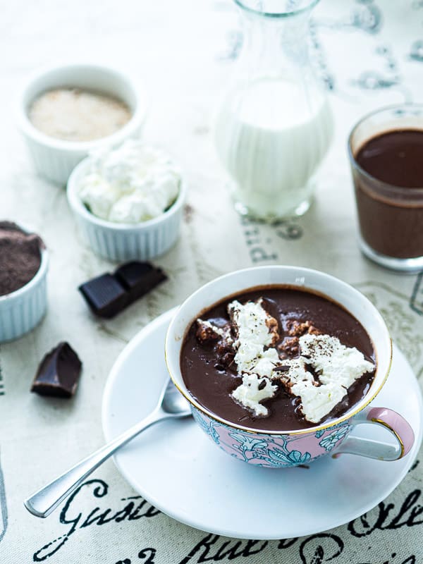 2 cups of hot chocolate next to whipped cream, sugar, milk, and cocoa powder.